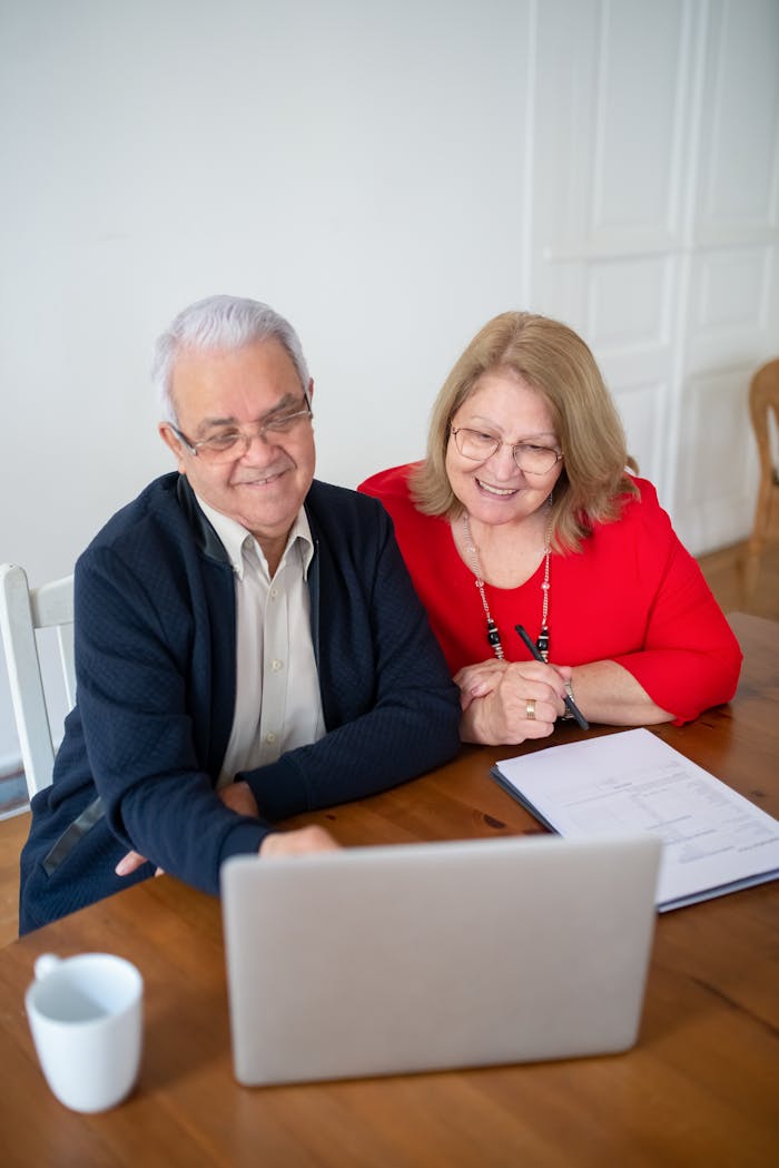 Senior couple sitting at a table, smiling while using a laptop indoors. Perfect for tech and lifestyle themes.